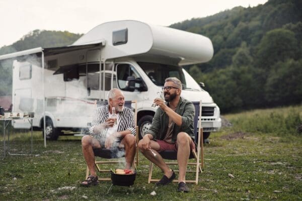 Two men sitting on camping chairs drinking beer and talking in front of RV - MitchMarket Finance